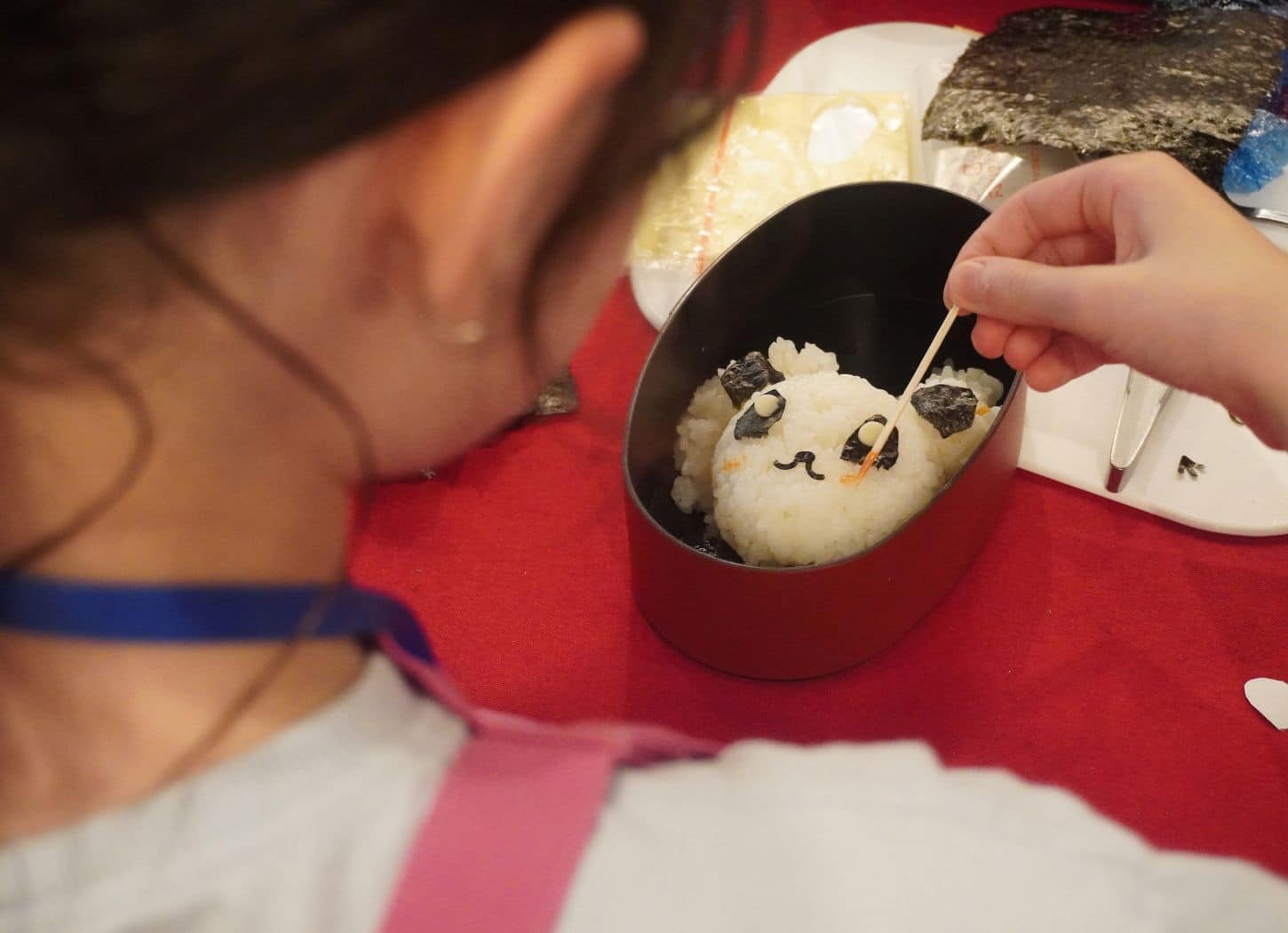 Child placing nori eyes on a panda rice ball in a bento box