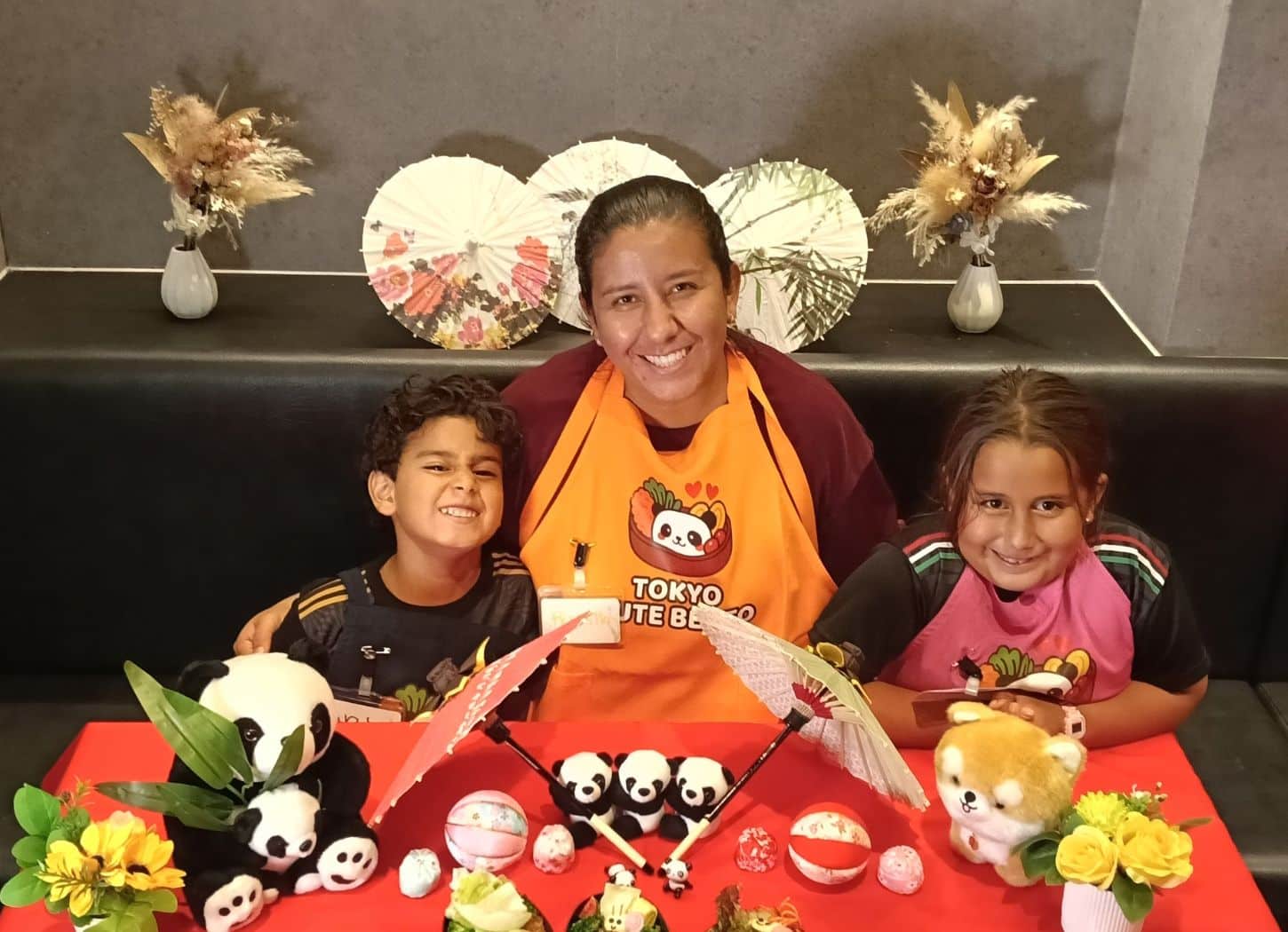 Parent and two children smiling behind panda plushies and paper parasols at Tokyo Cute Bento